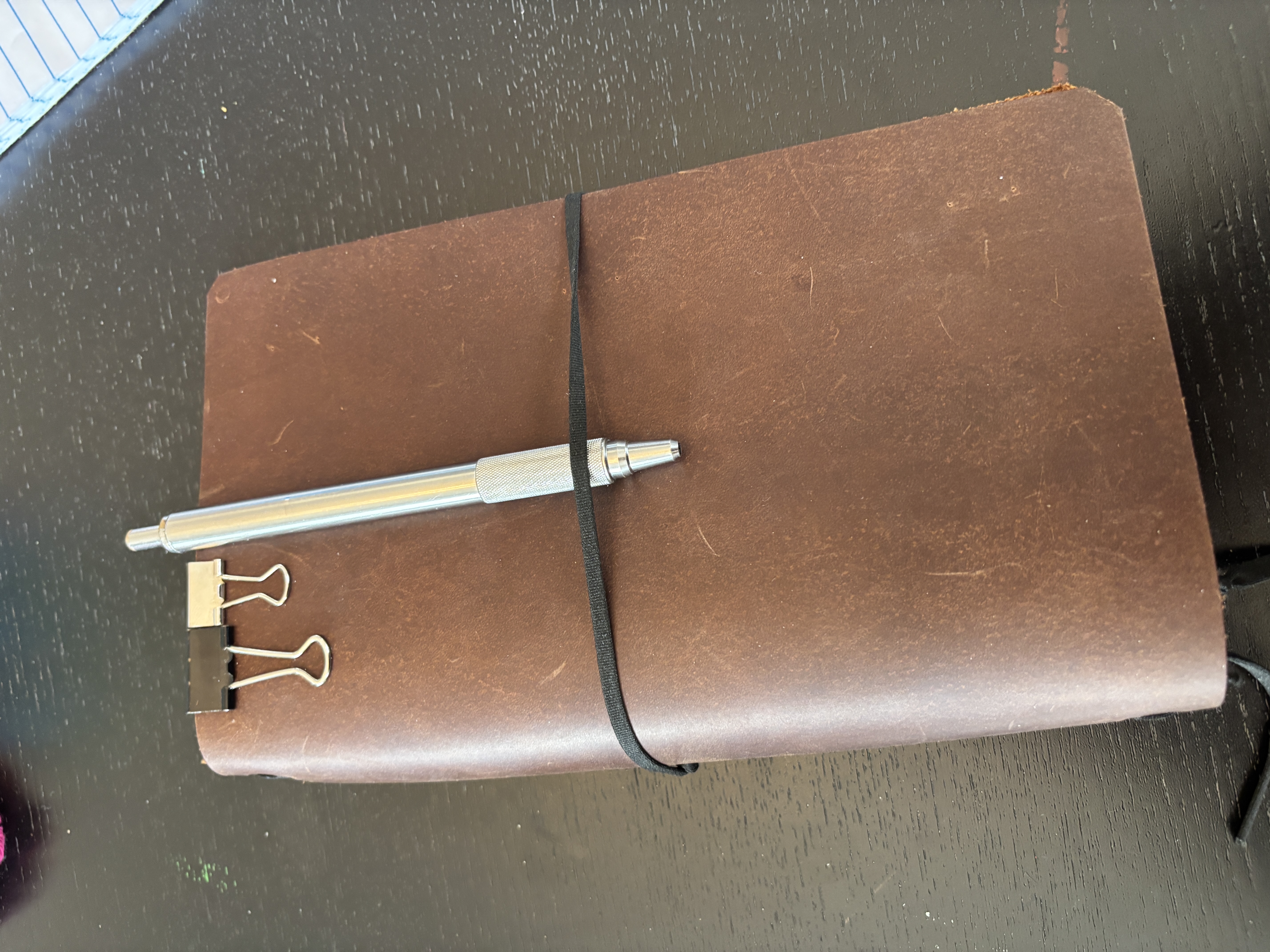 A leather notebook cover with an elastic band around it. A metal pen is clipped to the cover along with two binder clips.